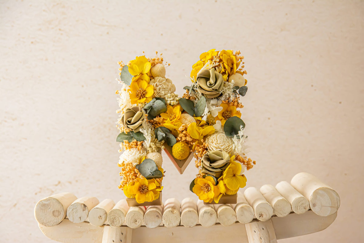 A dried flower letter M in yellow and sage on a wooden stool
