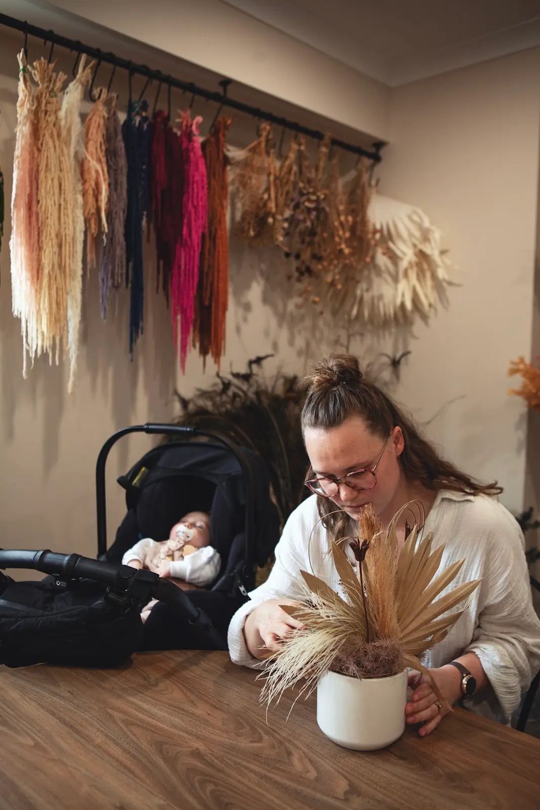 A mum arranges dried flowers in a vase with her baby in the pram beside her